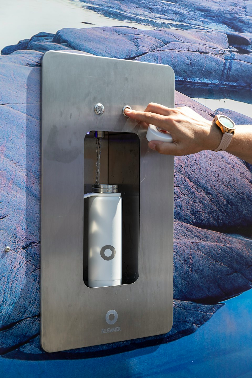 Hand pressing button on water dispenser to fill reusable bottle.